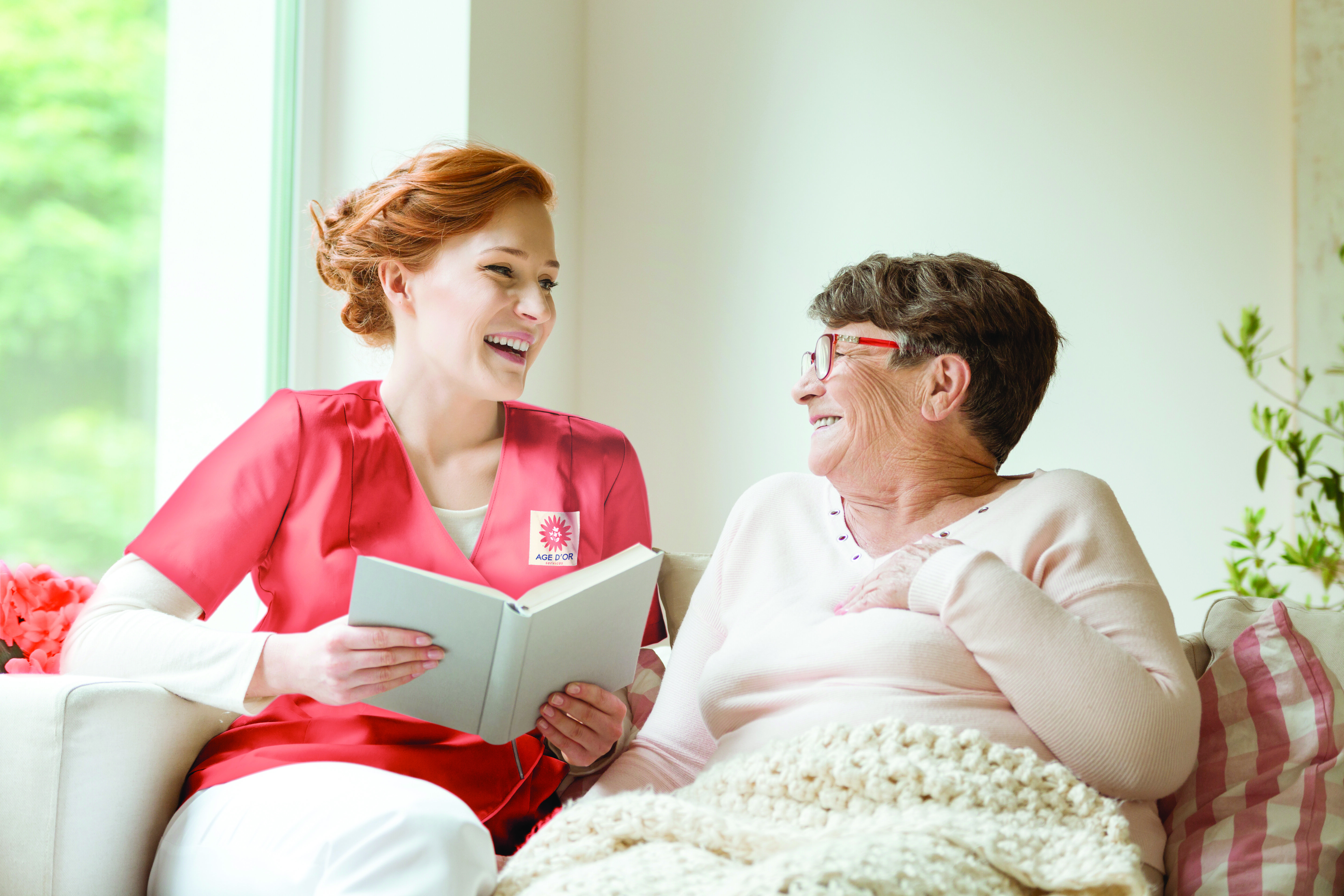 Photo d'illustration Axéo Services : Une infirmière à domicile est assise à côté d'une patiente dans son canapé. Elle est souriante. Soins à domicile, Parcours santé. Parcours de soin. Autonomie